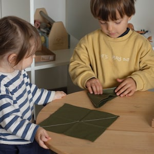 May include: Two children are sitting at a table, folding green fabric squares. The child on the left is wearing a striped shirt and the child on the right is wearing a yellow sweatshirt with the text "It's Play Out".