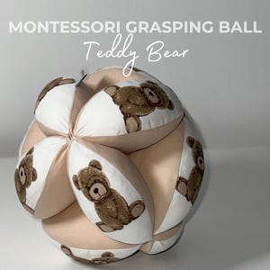 May include: A beige and white Montessori grasping ball with a teddy bear design. The ball is made of soft fabric and has six panels, each with a different teddy bear image.