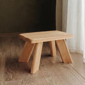 May include: A small, rectangular wooden stool featuring a light brown natural wood grain finish. The stool has a flat top surface with rounded edges and four sturdy, angled legs that provide a stable base for this functional furniture piece.