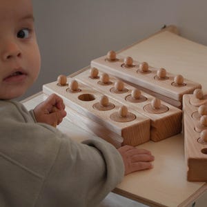 Peut inclure: Blocs de cylindres Montessori en bois avec des boutons. Les blocs sont brun clair et comportent un nombre variable de cylindres avec des boutons en bois. Les blocs sont disposés sur une surface en bois de couleur claire. Un enfant est au premier plan.
