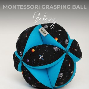 May include: A blue and black fabric grasping ball with a galaxy print. The ball has a tag that says "bawka" on it. The ball is a Montessori toy for babies and toddlers.