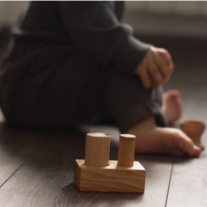 May include: A wooden toy with a rectangular base and two cylindrical pegs. The toy is made of light-colored wood and sits on a wooden floor. A child is in the background.