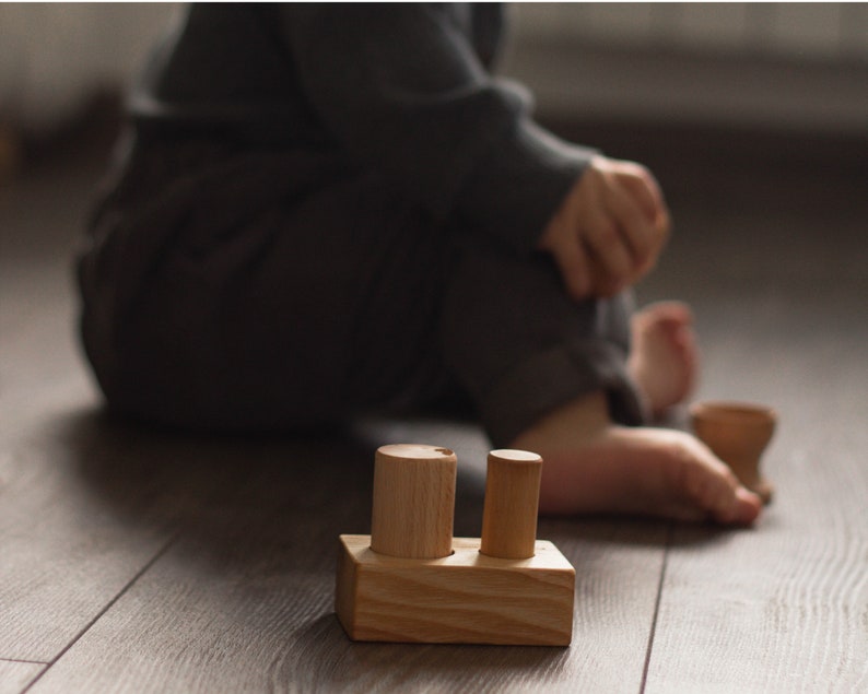 May include: A wooden toy with a rectangular base and two cylindrical pegs. The toy is made of light-colored wood and sits on a wooden floor. A child is in the background.