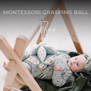 Puede incluir: Un bebé yace sobre una manta verde, sosteniendo una pelota de tela multicolor. La pelota está colgada de un gimnasio de juegos de madera con el texto "MONTESSORI GRASPING BALL" encima.