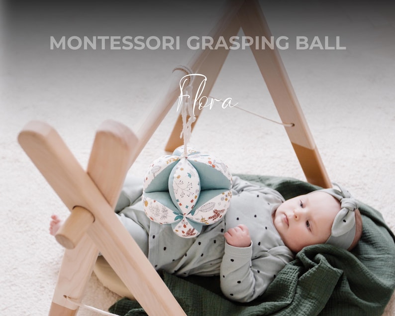 May include: A baby lies on a green blanket, holding a multi-colored fabric ball. The ball is hanging from a wooden play gym with the text "MONTESSORI GRASPING BALL" above it.