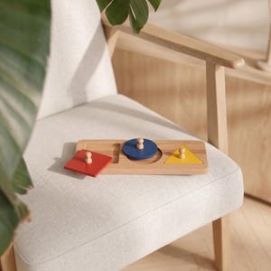 Op de afbeelding: Een houten vormensorteerbord met drie uitsparingen. De set bevat een rood vierkant, een blauwe cirkel en een gele driehoek, elk voorzien van een kleine houten knop om vast te pakken, rustend op een basis van lichtgekleurd hout.