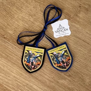 May include: Two shield-shaped religious pendants with the text "QUIS UT DEUS" and an illustration of a saint. One pendant has a black border, the other has a blue border. Both have blue and black ribbons attached. A white tag with the text "VATICAN Religious Gifts" is also present.