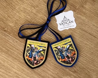 May include: Two shield-shaped religious pendants with the text "QUIS UT DEUS" and an illustration of a saint. One pendant has a black border, the other has a blue border. Both have blue and black ribbons attached. A white tag with the text "VATICAN Religious Gifts" is also present.
