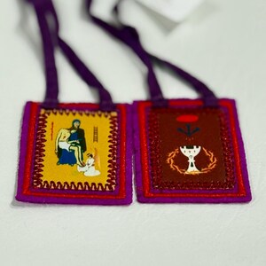 May include: Two religious scapulars with purple straps. One scapular features a yellow background with a religious image, the other has a brown background with a chalice and crown of thorns. Both are trimmed in red and purple.