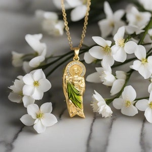 Puede incluir: Collar con colgante de oro con una imagen detallada de un santo con detalles en verde y dorado. El colgante está rodeado de pequeñas piedras transparentes. El collar se muestra sobre una superficie de mármol blanco con flores blancas.