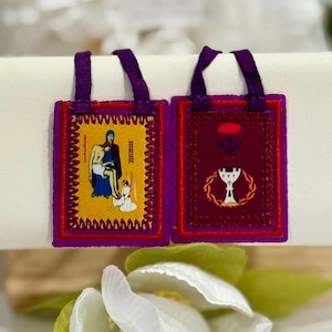 May include: Two rectangular religious pendants with purple borders and straps. One pendant has a yellow background with a religious illustration. The other has a burgundy background with a chalice and wreath design. Both are displayed on a white surface.