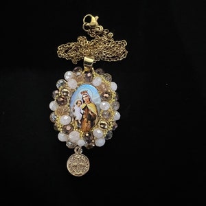 May include: Gold-toned necklace featuring an oval pendant with a religious image of a woman and child, surrounded by beads in shades of brown, white, and gold. A smaller, round charm hangs below. The necklace is set against a black background.