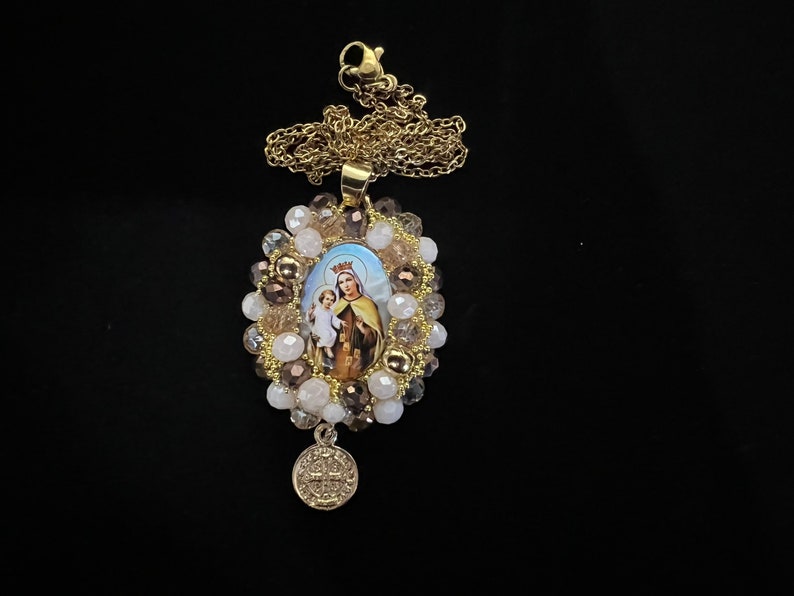 May include: Gold-toned necklace featuring an oval pendant with a religious image of a woman and child, surrounded by beads in shades of brown, white, and gold. A smaller, round charm hangs below. The necklace is set against a black background.