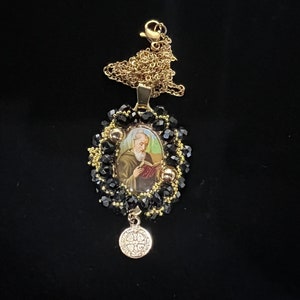 May include: A gold-toned necklace with a pendant featuring an image of a saint. The pendant is surrounded by black beads and gold accents. A smaller, round charm with text hangs below the pendant. The necklace is on a black background.