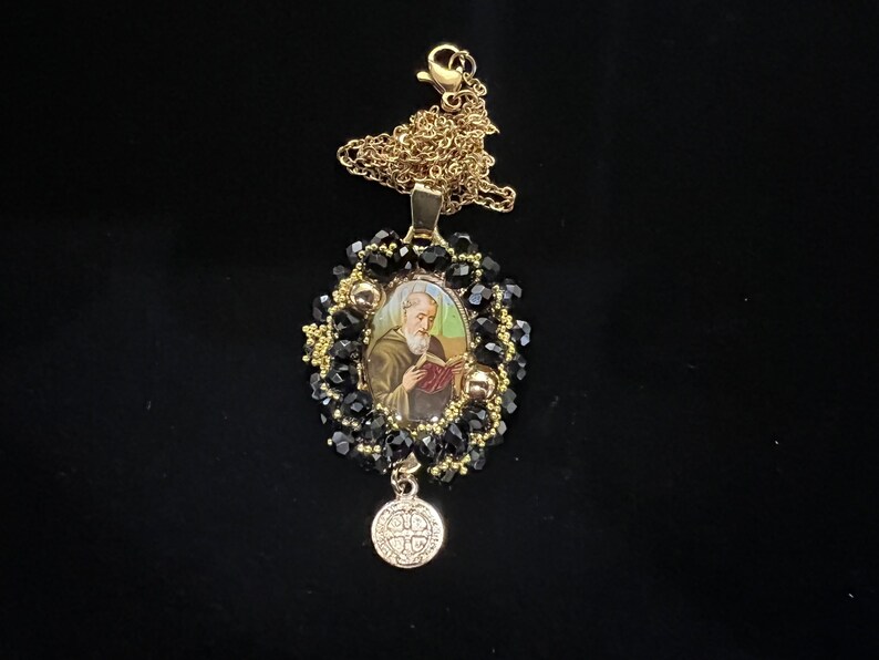 May include: A gold-toned necklace with a pendant featuring an image of a saint. The pendant is surrounded by black beads and gold accents. A smaller, round charm with text hangs below the pendant. The necklace is on a black background.