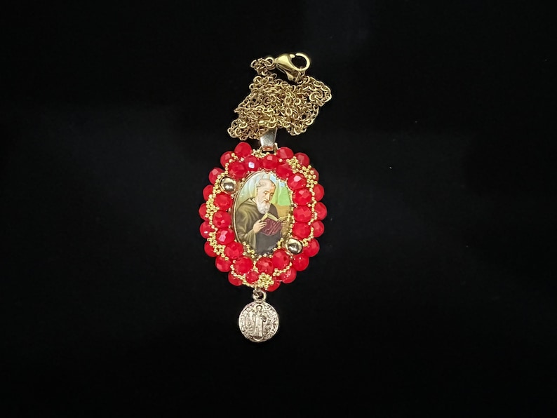 May include: A gold-toned necklace with a pendant featuring a religious image surrounded by red beads. A smaller, round charm with a religious emblem hangs below the pendant. The necklace is set against a black background.