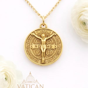 May include: Gold-toned pendant necklace featuring a detailed medallion with a central figure and surrounding text. The pendant is suspended from a delicate chain. The word "PAX" is visible on the top of the pendant. The word "VATICAN" is printed at the bottom.