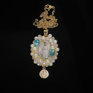May include: A gold-colored necklace with a pendant featuring an image of a religious figure. The pendant is surrounded by pink and white beads, with blue crystal accents. A smaller, round charm hangs below.