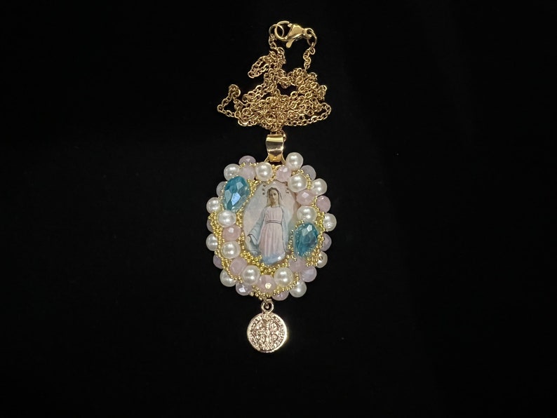 May include: A gold-colored necklace with a pendant featuring an image of a religious figure. The pendant is surrounded by pink and white beads, with blue crystal accents. A smaller, round charm hangs below.