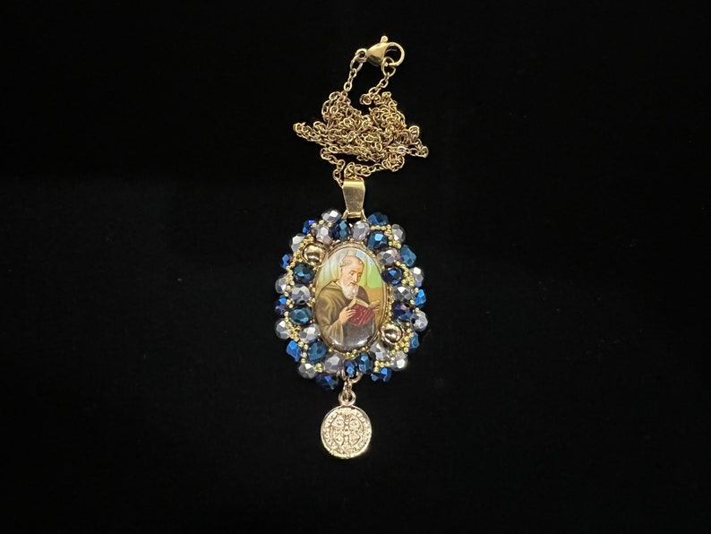May include: Gold-tone necklace featuring an oval pendant with a religious image, surrounded by blue and clear beads. A small, round charm dangles below. The chain and pendant have a vintage aesthetic.