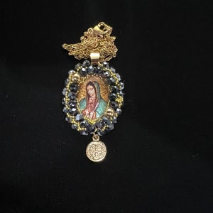 May include: Gold-toned necklace featuring a pendant with an image of the Virgin Mary. The pendant is surrounded by dark blue beads and gold accents, with a small round charm at the bottom. The chain is delicate and gold.