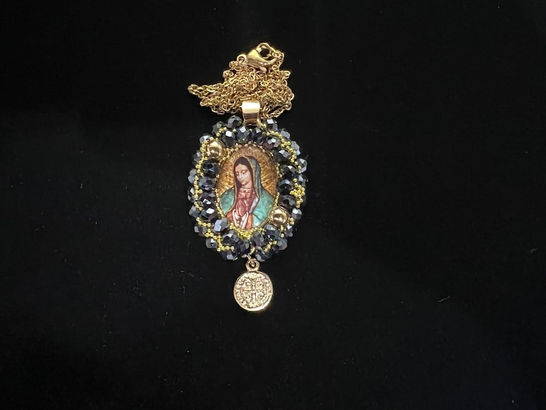 May include: Gold-toned necklace featuring a pendant with an image of the Virgin Mary. The pendant is surrounded by dark blue beads and gold accents, with a small round charm at the bottom. The chain is delicate and gold.