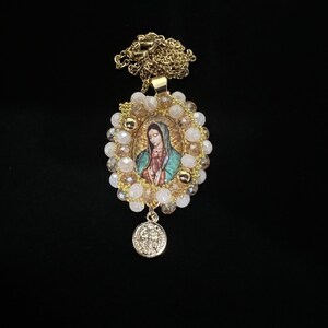May include: Gold-toned necklace featuring an oval pendant with an image of the Virgin Mary, surrounded by cream and gold beads. A smaller, round charm hangs below. The chain and pendant have a shiny finish, set against a black background.