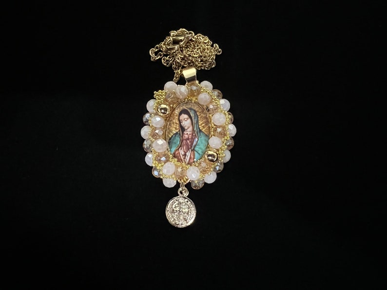 May include: Gold-toned necklace featuring an oval pendant with an image of the Virgin Mary, surrounded by cream and gold beads. A smaller, round charm hangs below. The chain and pendant have a shiny finish, set against a black background.