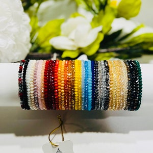 Puede incluir: Una colección de pulseras de cuentas de varios colores, incluyendo rojo, naranja, amarillo, azul y verde. Las pulseras se exhiben sobre una superficie blanca, con una etiqueta "Vatican Religious Gifts."
