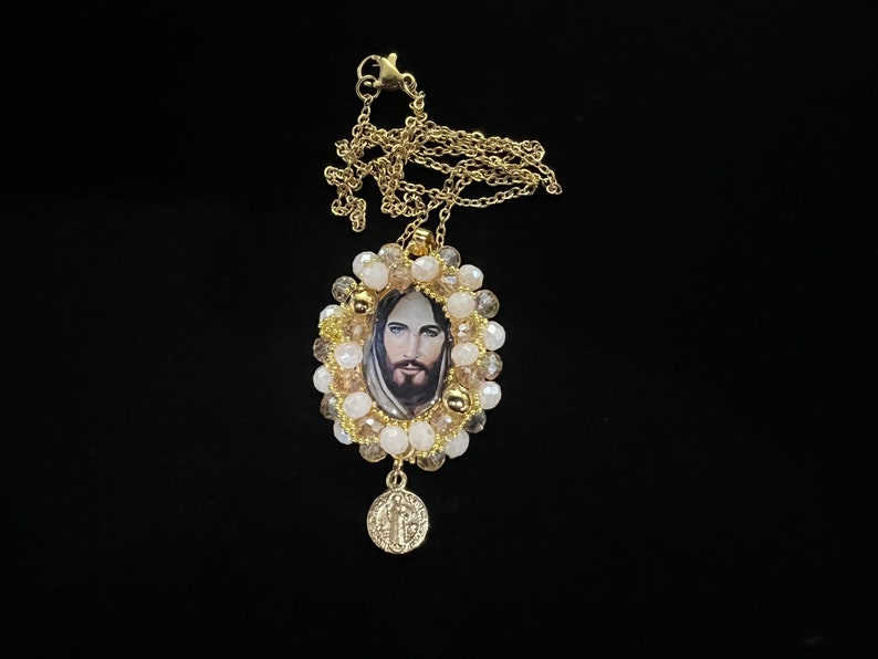 May include: Gold-toned necklace featuring a pendant with an image of Jesus Christ. The pendant is surrounded by cream and clear beads. A smaller, round charm with religious text hangs below the pendant. The necklace is displayed against a black background.