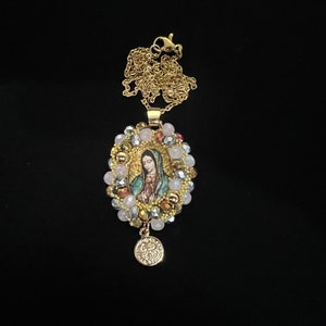 May include: Gold-toned necklace featuring a pendant with an image of the Virgin Mary. The pendant is surrounded by a halo of pink, clear, and gold beads. A small, round charm hangs below the pendant.
