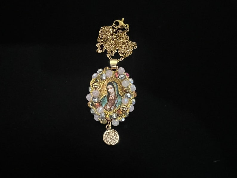 May include: Gold-toned necklace featuring a pendant with an image of the Virgin Mary. The pendant is surrounded by a halo of pink, clear, and gold beads. A small, round charm hangs below the pendant.