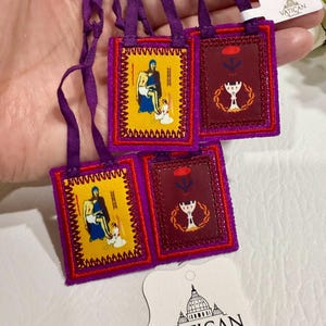 May include: A collection of small, embroidered religious items with purple straps. The items feature images of religious figures and symbols in yellow and burgundy. The tag reads "VATICAN Religious Gifts".