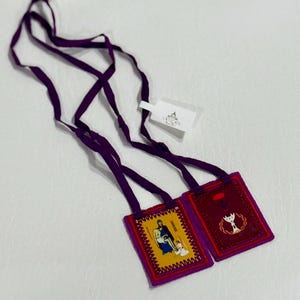 May include: Two religious scapulars with purple straps and tags from the Vatican. One scapular has a yellow background with a religious image, and the other has a red background with a religious symbol.