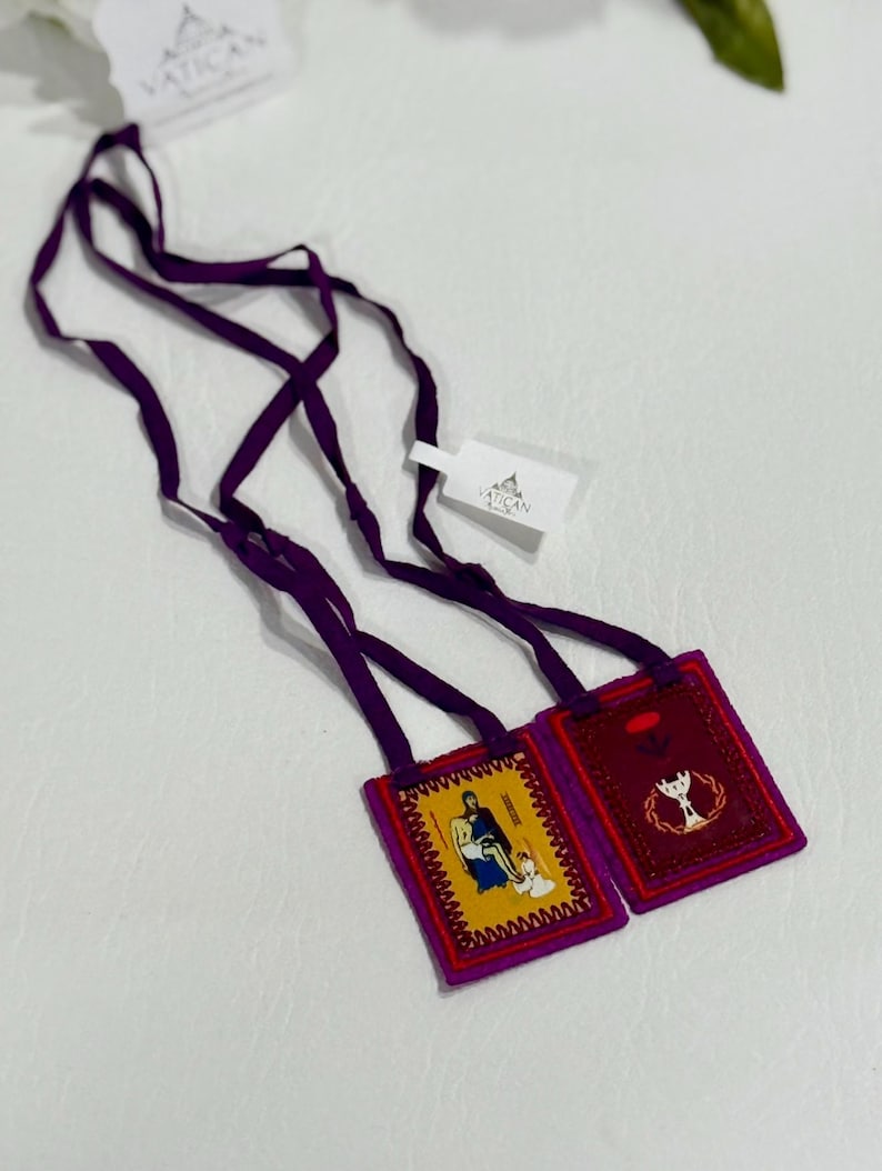 May include: Two religious scapulars with purple straps and tags from the Vatican. One scapular has a yellow background with a religious image, and the other has a red background with a religious symbol.