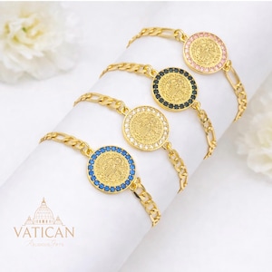 May include: Gold-toned bracelets with circular pendants. Each pendant features a religious emblem and is surrounded by colored gemstones: pink, black, white, and blue. The bracelets have a chain link design. The Vatican Religious Gifts logo is visible.