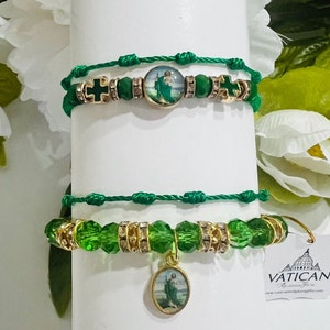 Puede incluir: Dos pulseras verdes y doradas con dijes religiosos. La pulsera superior tiene un dije redondo con una imagen de un santo. La pulsera inferior tiene un dije redondo con una imagen de un santo y cuentas de vidrio verdes.