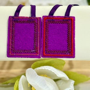 May include: Two rectangular purple felt ornaments with red and purple embroidered borders and purple ribbon loops. The ornaments are displayed on a cream-colored surface, with a white and green flower in the foreground.