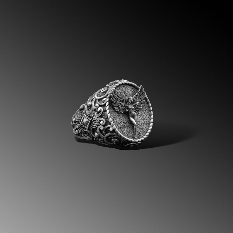 Puede incluir: Un anillo de plata con un dise&ntilde;o ovalado que presenta una imagen esculpida detallada de un &aacute;ngel con alas. La banda del anillo est&aacute; intrincadamente dise&ntilde;ada con patrones florales.