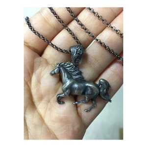 Puede incluir: Un collar de plata con un colgante de caballo con un dise&ntilde;o detallado. El caballo est&aacute; corriendo y tiene una melena y una cola fluidas. El collar est&aacute; en una cadena negra.