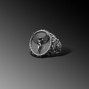 Puede incluir: Un anillo de plata con un dise&ntilde;o ornamentado que presenta una figura alada en el centro.