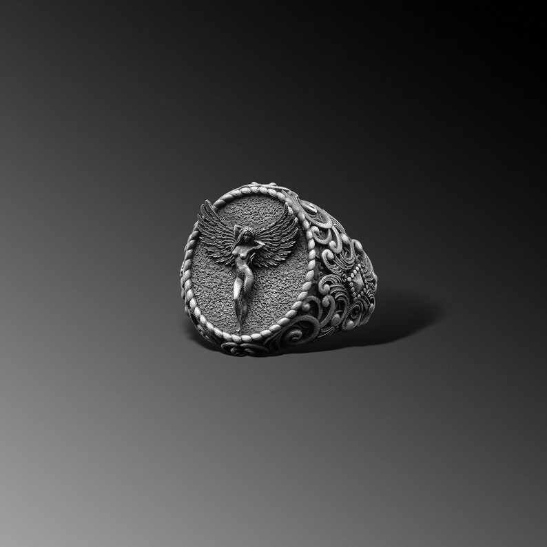 Puede incluir: Un anillo de plata con un dise&ntilde;o ornamentado que presenta una figura alada en el centro.
