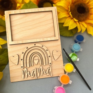 May include: Wooden craft kit featuring a rainbow and the word "mama" with a picture frame at the top. The kit includes paint pots in various colors and a paintbrush.