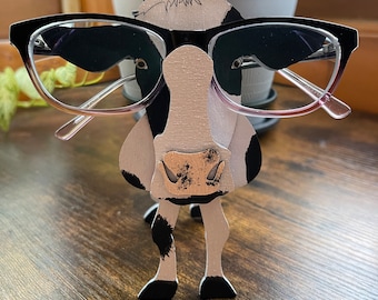 May include: A black and white cow-shaped wooden glasses holder with black eyes and a pink nose. The holder is standing on a wooden surface.