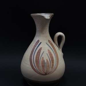 Puede incluir: Una jarra de cerámica con base de color crema y un motivo floral pintado a mano en tonos marrones en la parte frontal. El recipiente presenta un cuello estrecho, un borde ensanchado con pico vertedor y una asa curva en el lado derecho.