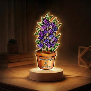 May include: A decorative LED lamp featuring a potted plant with purple flowers and green leaves. The pot is brown and has the text "Forever grateful for your love". The lamp sits on a wooden base and emits a warm glow.