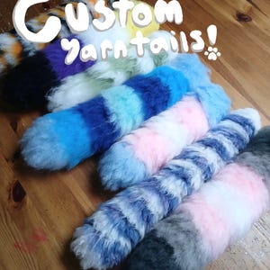 May include: A collection of fluffy, multi-colored yarn tails in various patterns and color combinations, including blue, pink, white, grey, and orange. The text 'Custom yarntails!' is overlaid in white on the image of these soft, textured accessories.