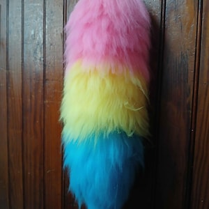 May include: A long, plush, faux fur tail accessory featuring a tri-color design. The top section is bright pink, the middle section is vibrant yellow, and the bottom section is a saturated cyan blue. The fur is thick, textured, and layered.