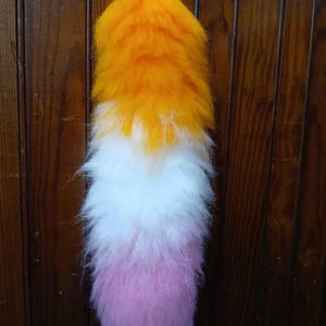 May include: A fluffy, tri-color tail prop, featuring a gradient of orange, white, and pink. The tail is made of soft, textured material and is suspended by a metal wire. The prop is designed for cosplay or costume use.