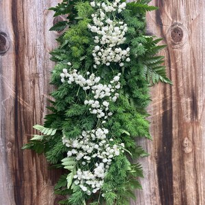 以下が含まれることがあります： 木製の壁に白いカスミソウの花と緑のシダの長いガーランドがぶら下がっている